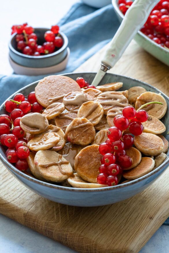 Bowl of mini pancakes drizzled with syrup, topped with fresh red currants on a wooden board.