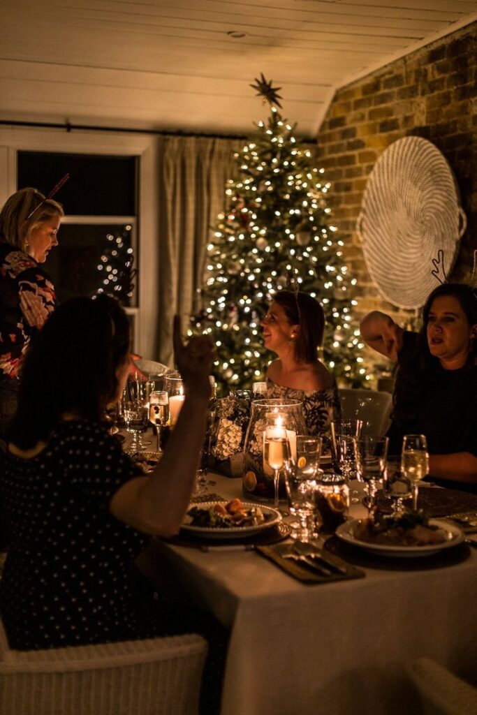 Cozy holiday dinner with friends around a festive table and a decorated Christmas tree in the background.