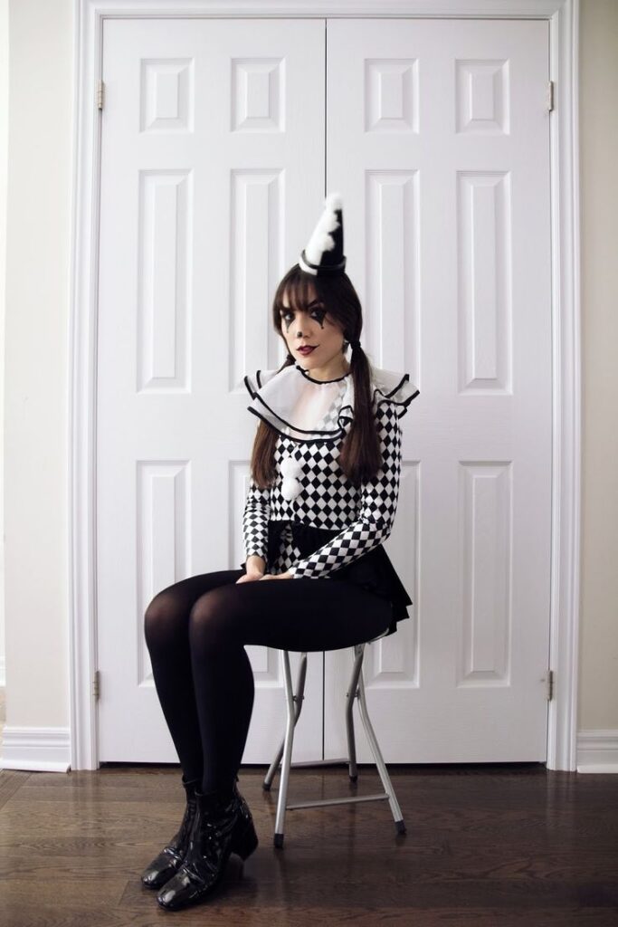 Person in black and white harlequin costume with cone hat sitting on chair, white doors in background.