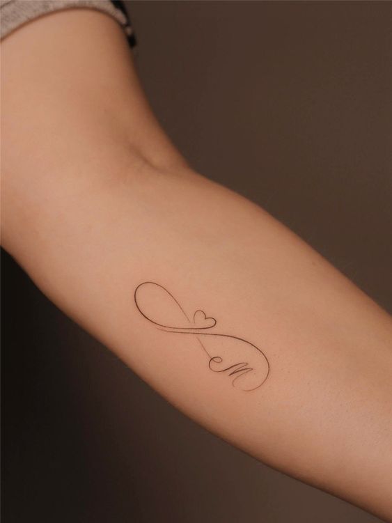 Minimalist infinity tattoo with a heart and letter 'M' on forearm, symbolizing eternal love and connection.