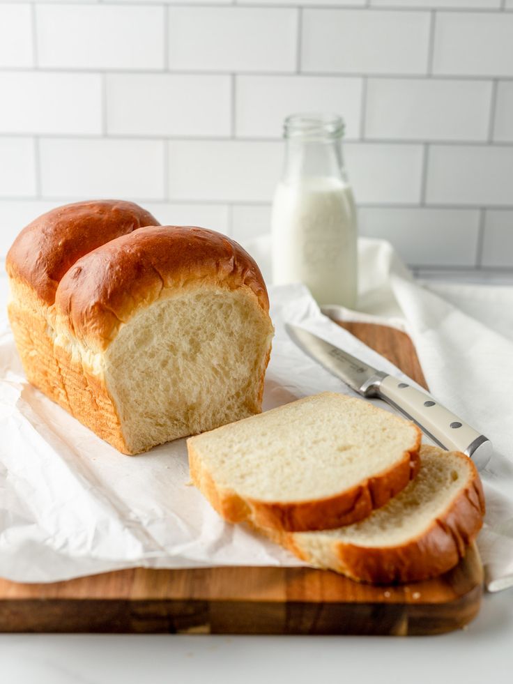 Freshly baked bread with slices on a wooden board, accompanied by a knife and a bottle of milk in the background.