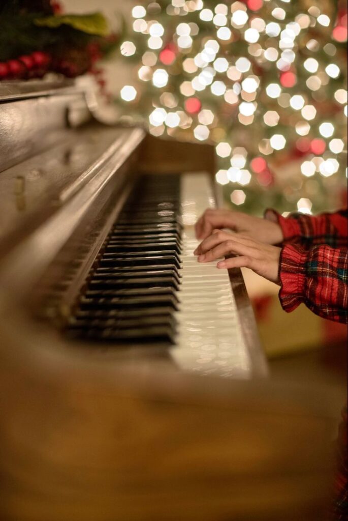 Hands playing a piano with festive Christmas tree lights in the background.