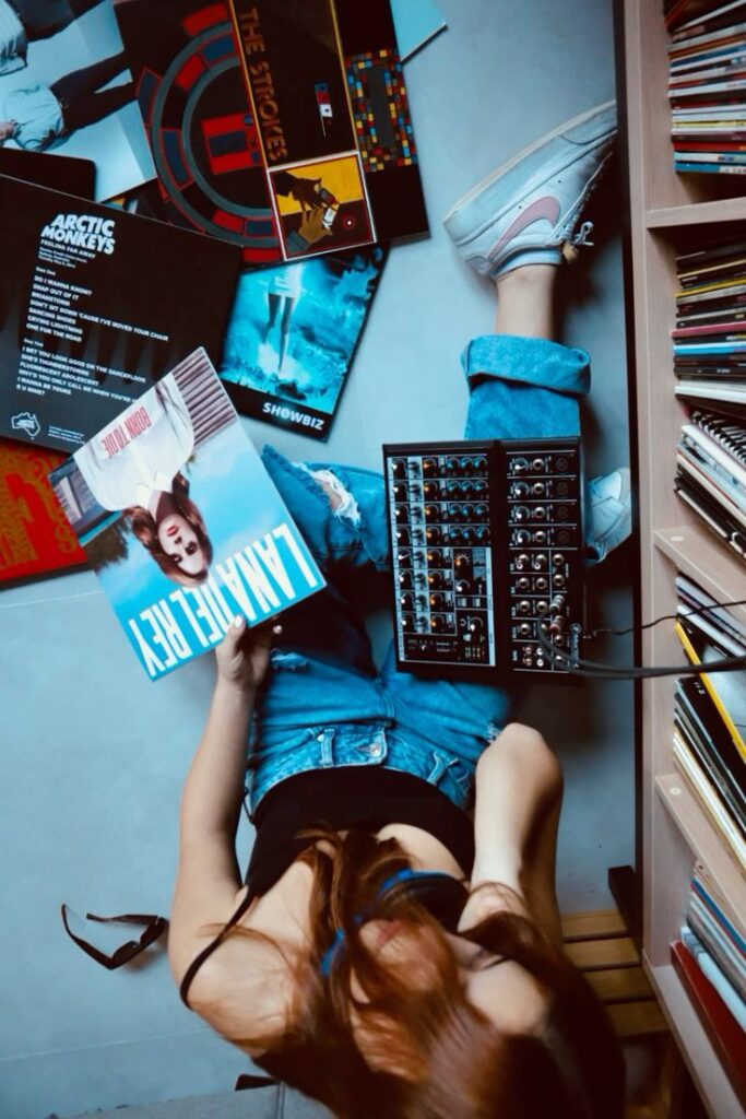 Person listening to vinyl records with a sound mixer on the floor, surrounded by albums and wearing casual jeans.