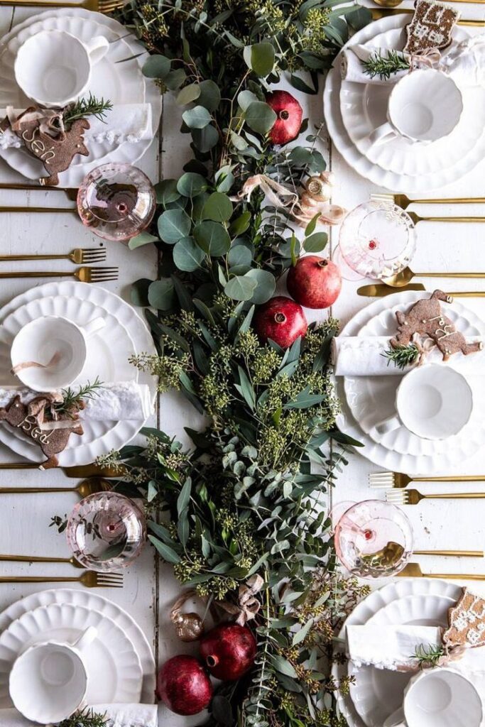 Elegant holiday table setting with greenery, pomegranates, gold cutlery, and white plates, creating a festive atmosphere.