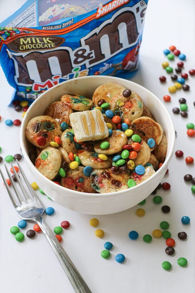 Mini pancakes topped with M&Ms, butter, and syrup in a bowl, next to a pack of M&M's Minis.