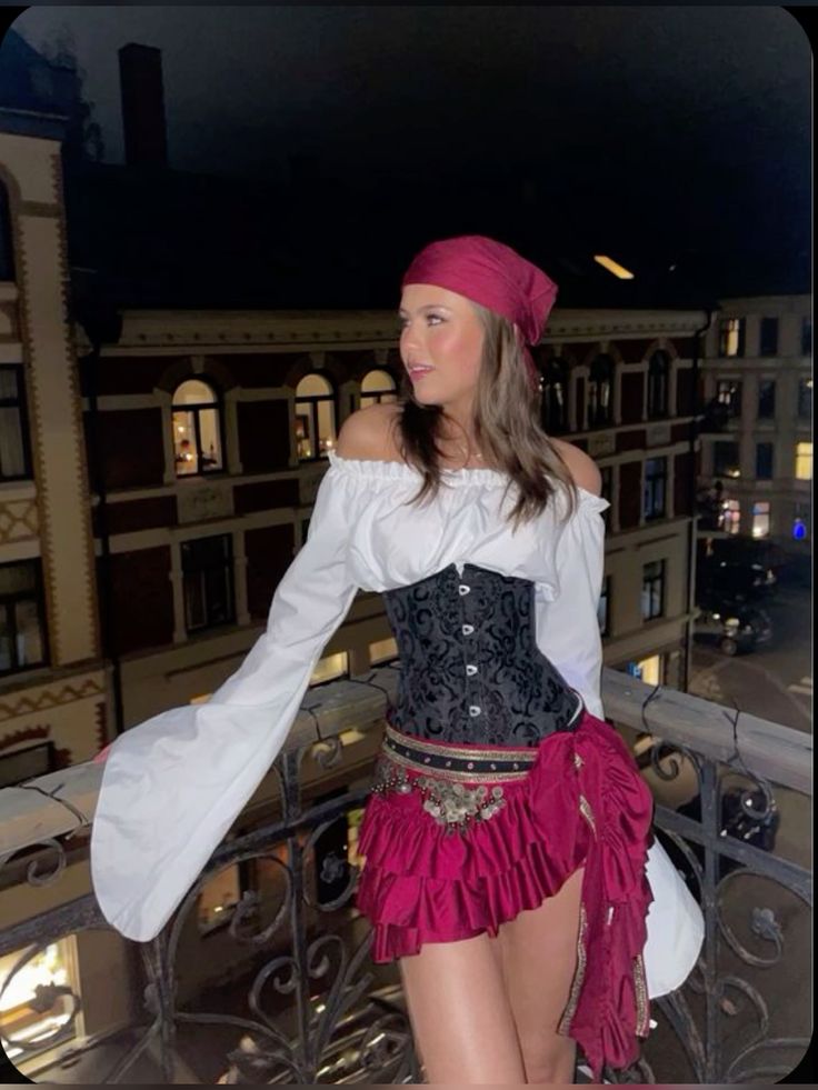 Woman in pirate costume on a balcony at night, wearing a white blouse, corset, and red skirt with matching headscarf.