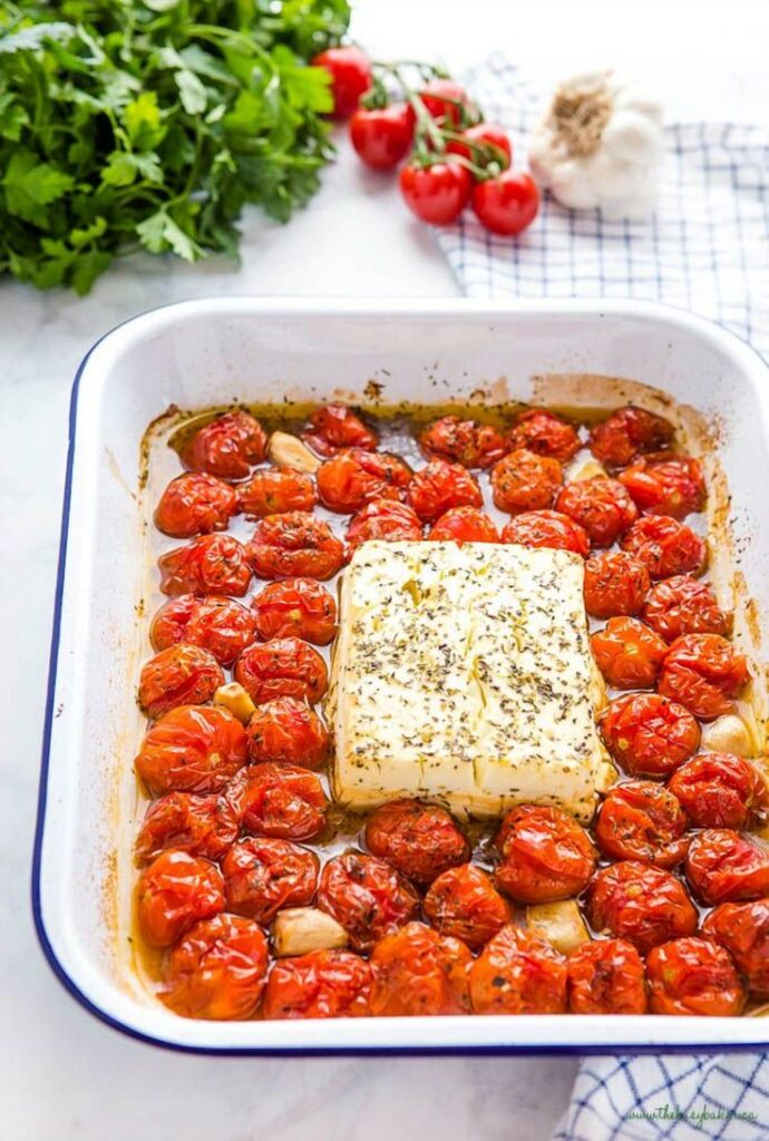 Baked feta with cherry tomatoes, garlic, and herbs in a white dish, garnished with fresh parsley and vine tomatoes.