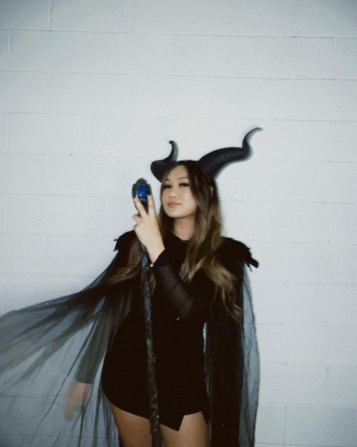 Person in a black costume with horned headpiece holding a staff, against a white brick wall.