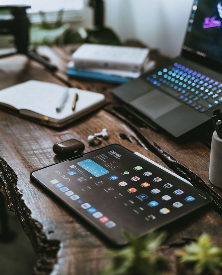 Tablet with home screen apps on a wooden desk, near an open laptop, notebook, and wireless earbuds.