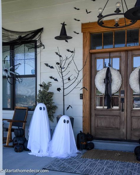 Halloween-themed porch with ghost decorations, bats, spider webs, and wreaths on a wooden door.