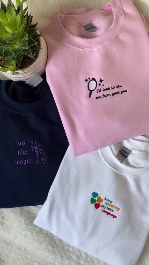 Three colorful embroidered shirts with unique designs, next to a potted succulent plant on a cozy surface.