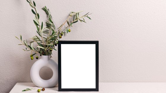 Minimalist decor with a black picture frame and olive branches in a white vase against a neutral background.