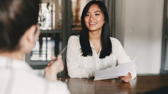 Woman in a white blouse smiling during a job interview, holding papers across a table from the interviewer.