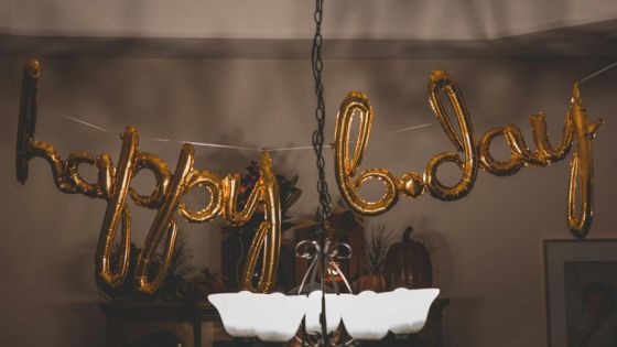 Gold balloon letters spelling happy bday hang above a light fixture at a birthday celebration indoors.