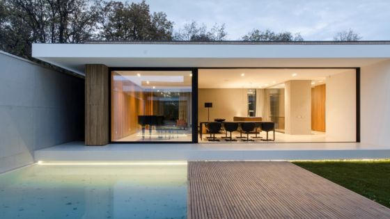 Modern minimalist house with large glass windows, pool, and wooden deck, set amid lush greenery.