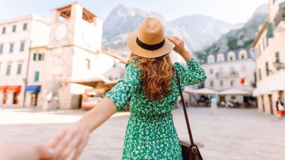 Woman in green dress holding hand, exploring an old European town square with mountain backdrop. Travel and adventure theme.