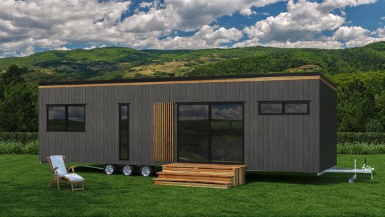 Modern mobile tiny house on grassy field with mountain backdrop under a partly cloudy sky.