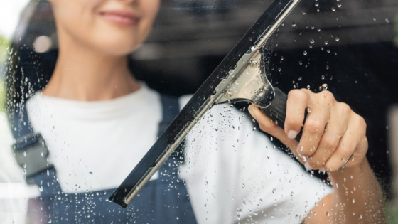 Person using a squeegee to clean a wet window, wearing a white shirt and overalls.