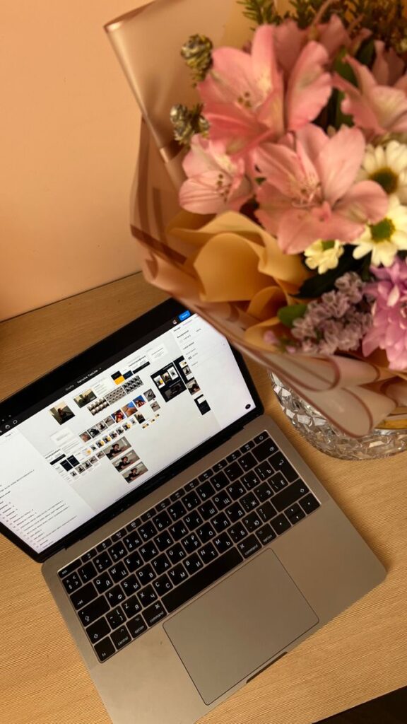 Open laptop with image gallery on screen beside a bouquet of pink and white flowers on a wooden desk.