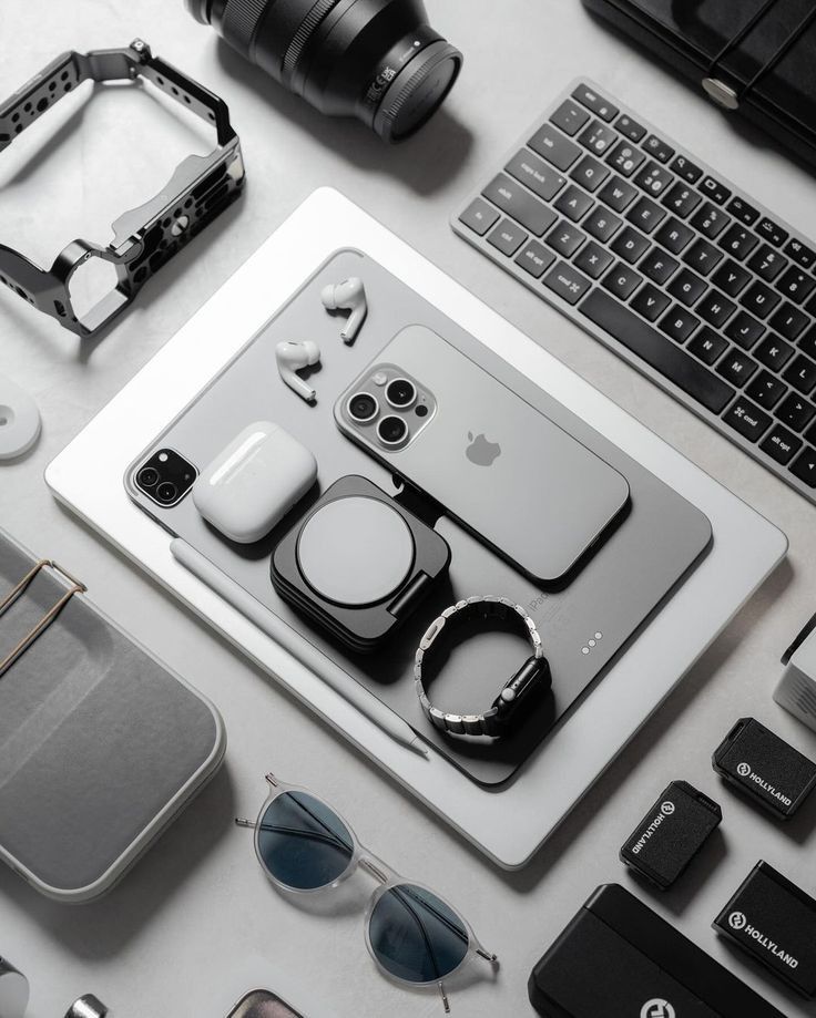 Modern tech gadgets neatly arranged: smartphone, earbuds, keyboard, camera lens, smartwatch, tablet, sunglasses.