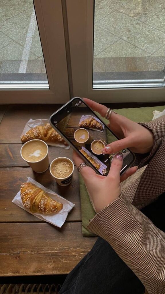 Person photographing coffee and croissants on a wooden table near a window. Cozy morning cafe scene.