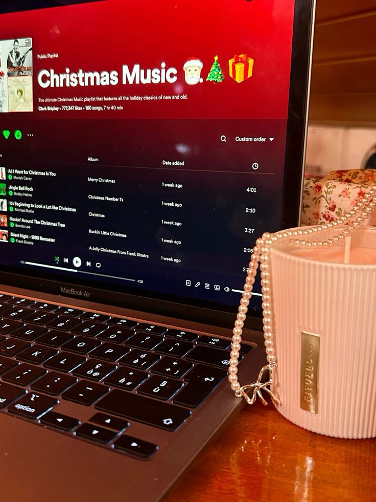 Laptop playing Christmas music on Spotify next to a pink candle and pearl necklace.