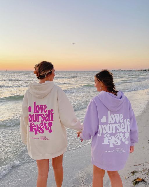 Two people in Love Yourself First hoodies hold hands on a beach at sunset, symbolizing self-love and friendship.