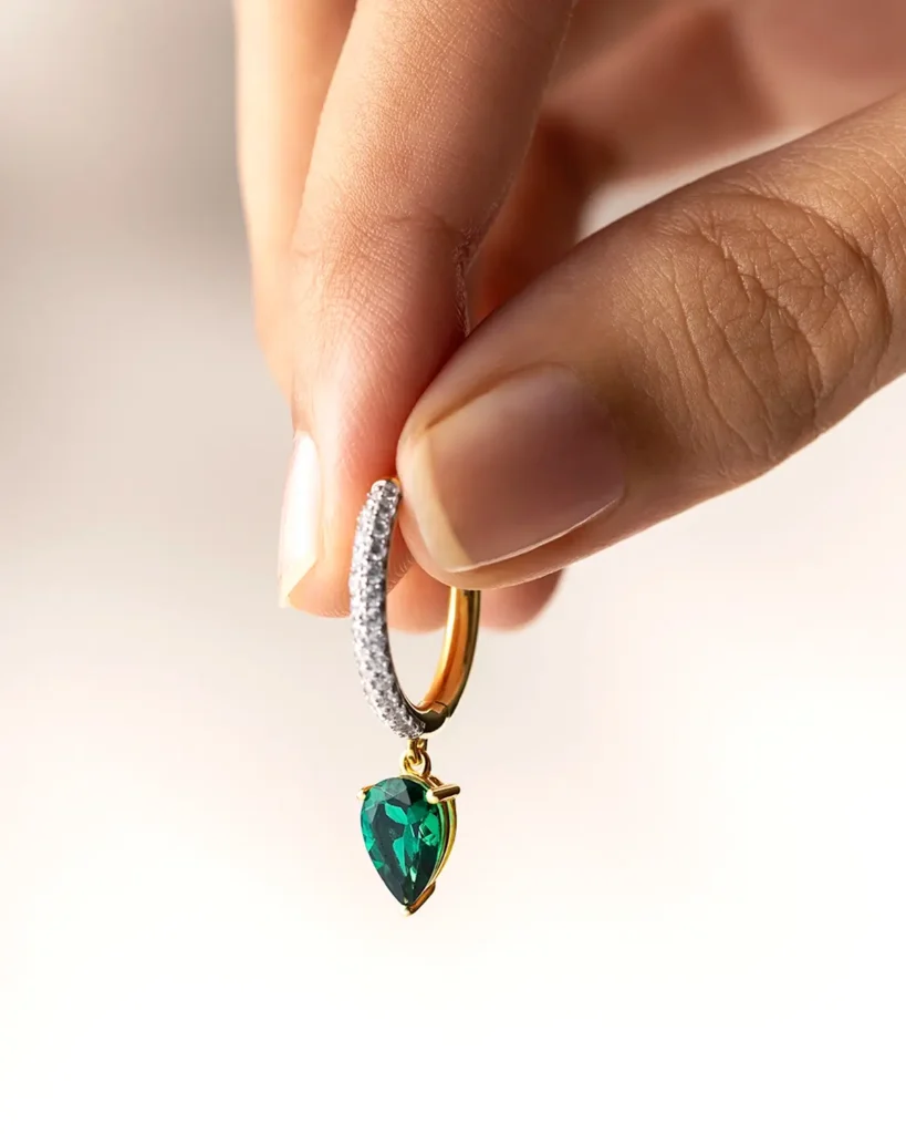 Hand holding a gold hoop earring with a green gemstone teardrop pendant against a neutral background.