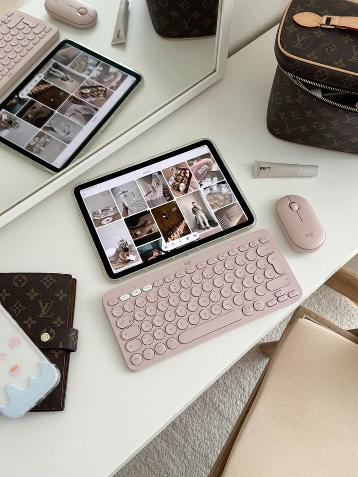 Stylish workspace with tablet, pink keyboard, and mouse on a white desk next to a mirror and designer bag.