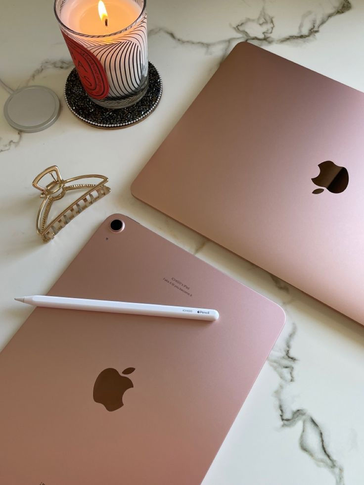 Rose gold Apple devices with a candle and hair clip on marble surface, featuring an Apple Pencil for creative workspace.