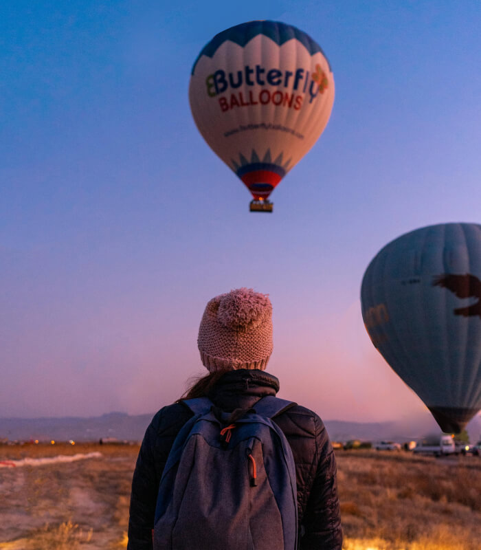 Person watching hot air balloons during sunrise, wearing a backpack and warm hat, in an open landscape.