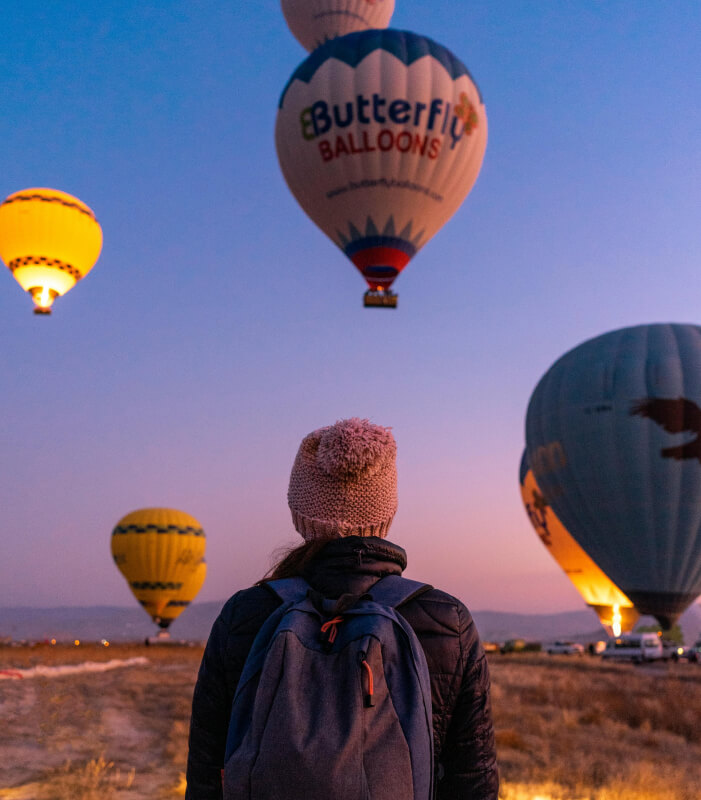 Person watching hot air balloons at sunrise, wearing a backpack and beanie in a scenic landscape.