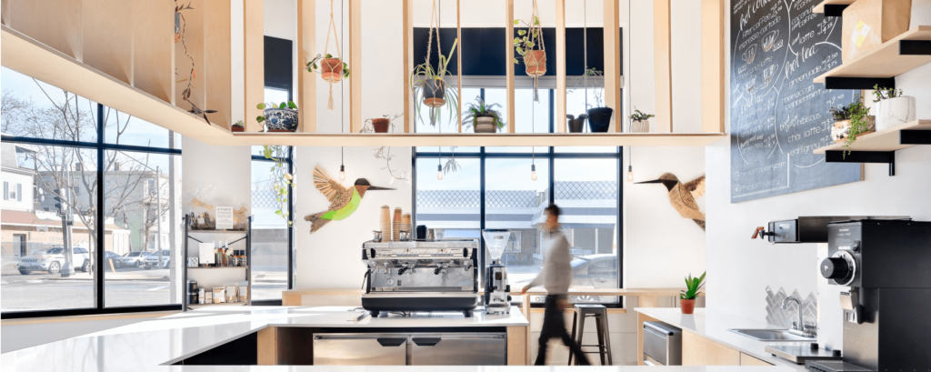 Modern cafe interior with plants, coffee machines, and menu board, featuring natural light and decorative wall art.