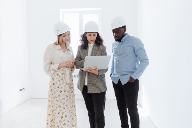 Three architects in hard hats discussing a project while reviewing plans on a laptop in a bright, unfinished room.