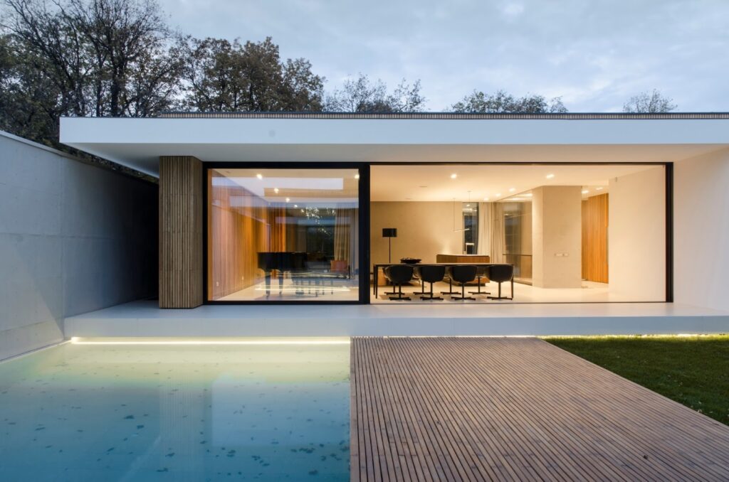 Modern home exterior with large windows, wooden deck, and pool, showcasing sleek minimalist design.