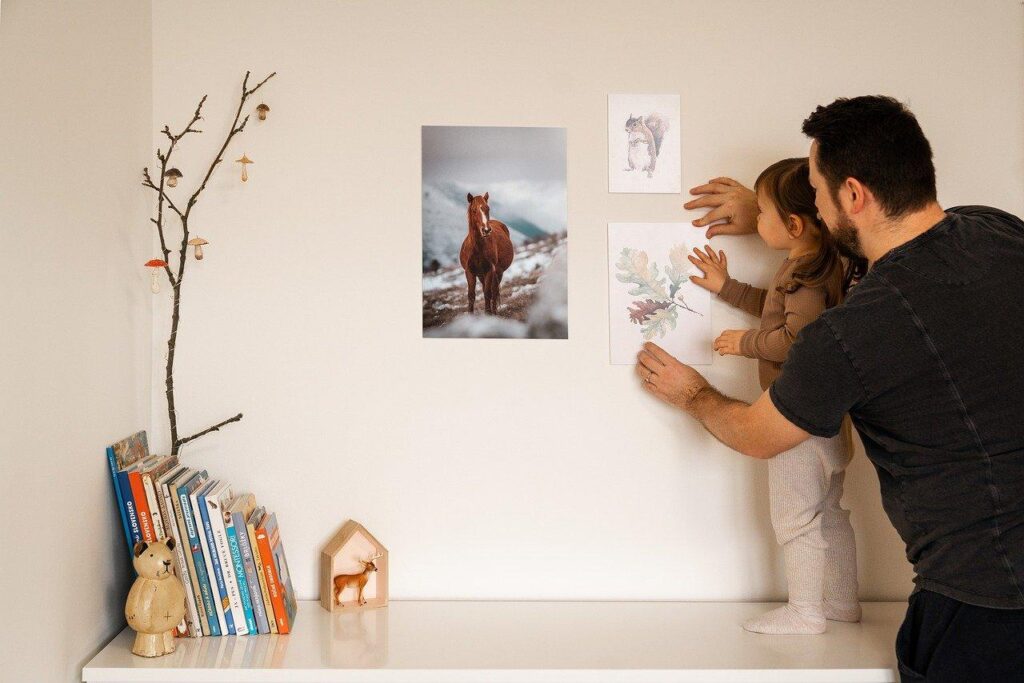 Father and child hang artwork above a shelf with books and decorations in a cozy room.