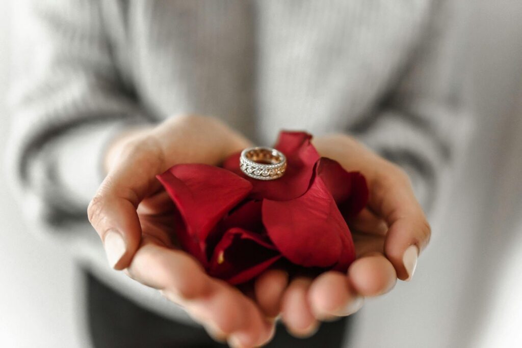 Hands holding red rose petals and a diamond ring, symbolizing love and engagement.