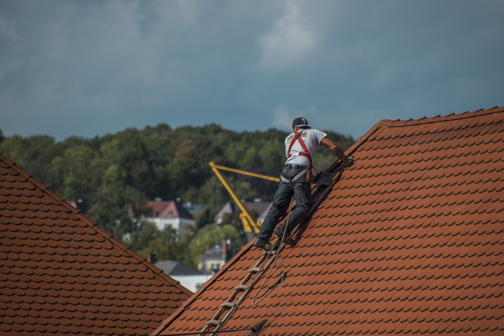 Roofer working on a steep red tile roof with harness and ladder, against a backdrop of trees and cloudy sky.