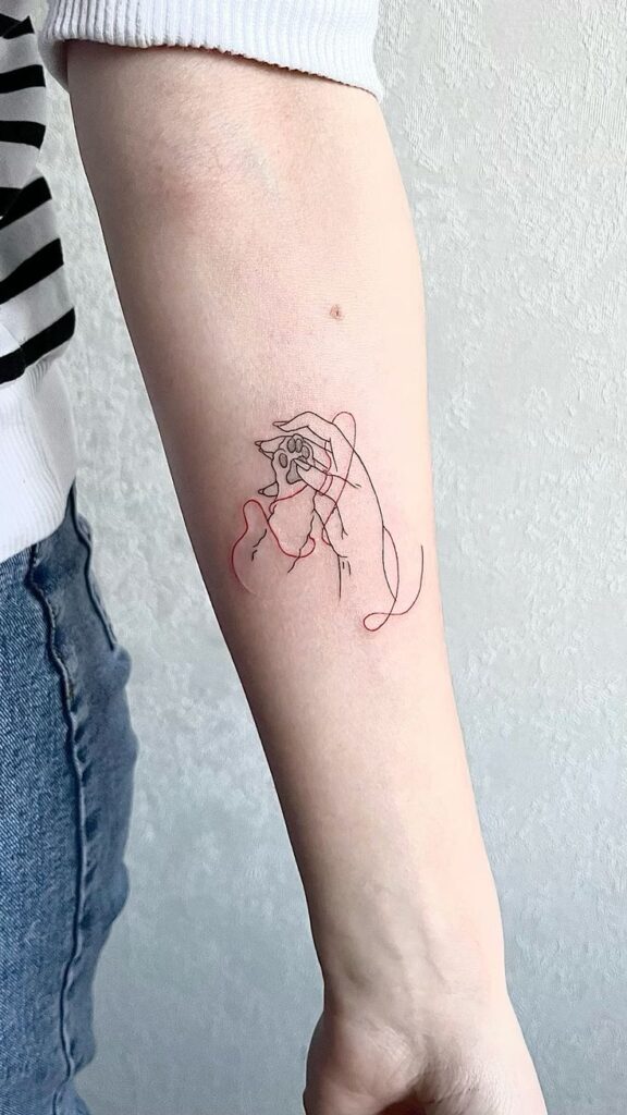 Minimalist line art tattoo of hands and a paw with a red thread on forearm, denim jeans and striped shirt sleeve visible.