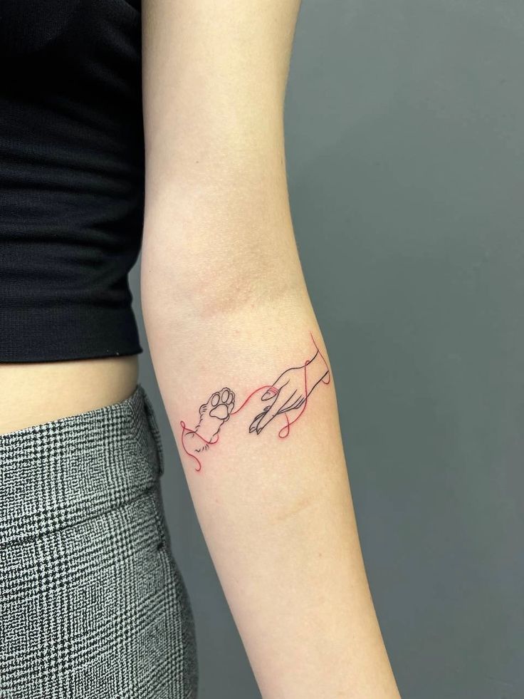 Minimalist tattoo of hand and paw connected by a red thread on forearm.