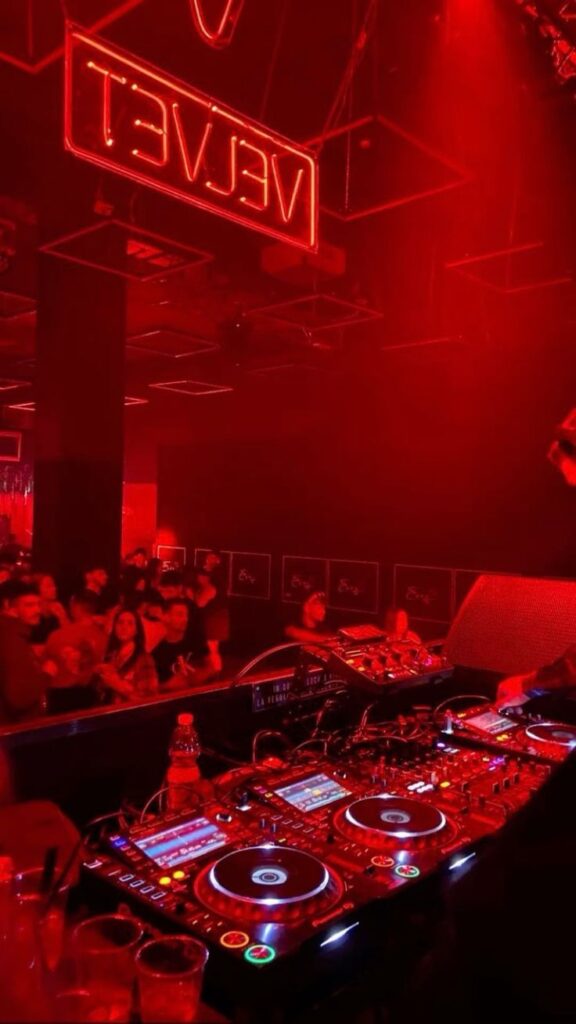 DJ booth with vibrant red lighting and crowd at a club, neon “Velvet” sign overhead.