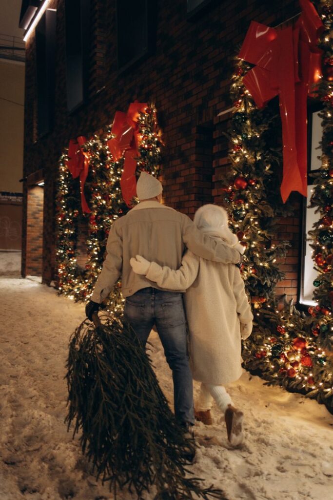 Зимняя фотосессия пара Couple walking with a Christmas tree in snowy street, festive lights and decorations on brick wall. Cozy winter scene. | Sky Rye Design Couple walking with a Christmas tree in snowy street, festive lights and decorations on brick wall. Cozy winter scene.