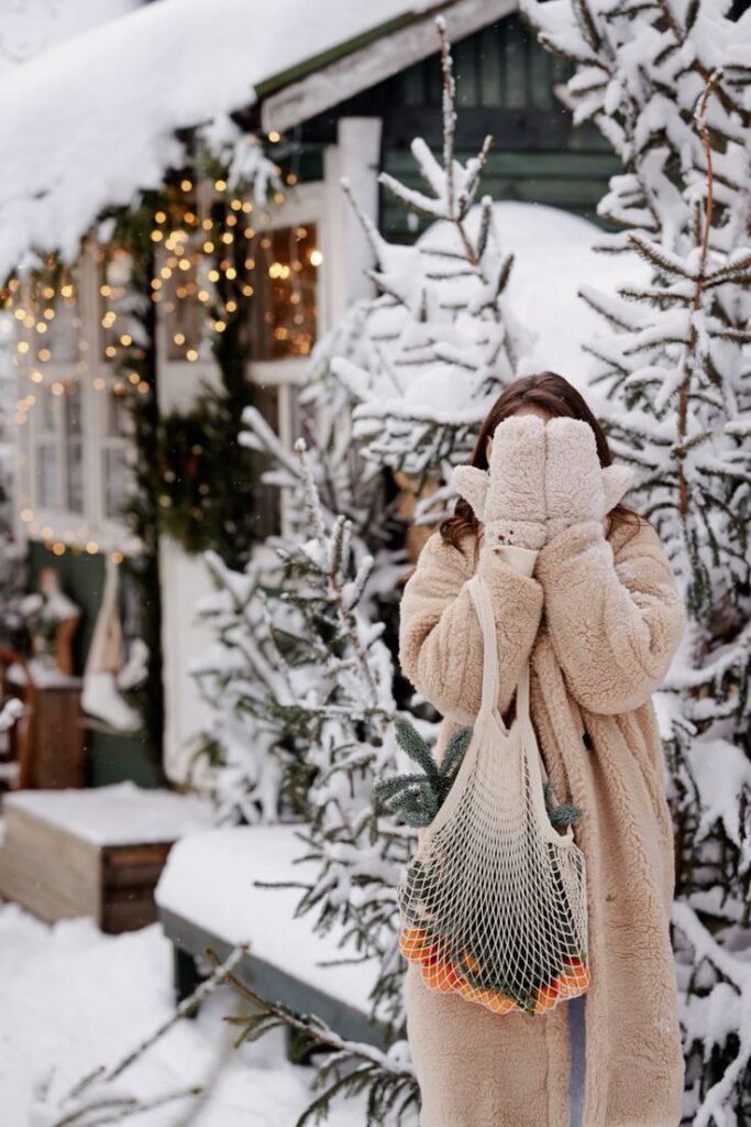 Зимняя фотосессия Cozy winter scene: woman in fur coat hides face, holding produce bag near snowy cabin with festive lights. | Sky Rye Design Cozy winter scene: woman in fur coat hides face, holding produce bag near snowy cabin with festive lights.