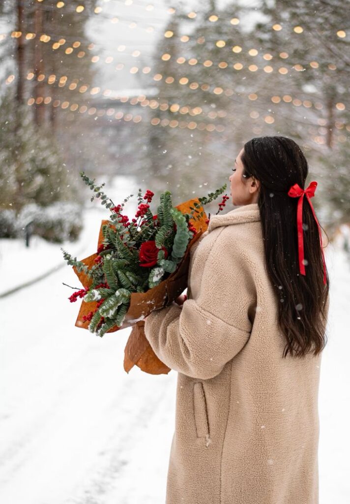 ФОТОСЕССИЯ ЗИМОЙ Woman in winter coat holding Christmas bouquet, standing in snowy landscape with festive lights. | Sky Rye Design Woman in winter coat holding Christmas bouquet, standing in snowy landscape with festive lights.
