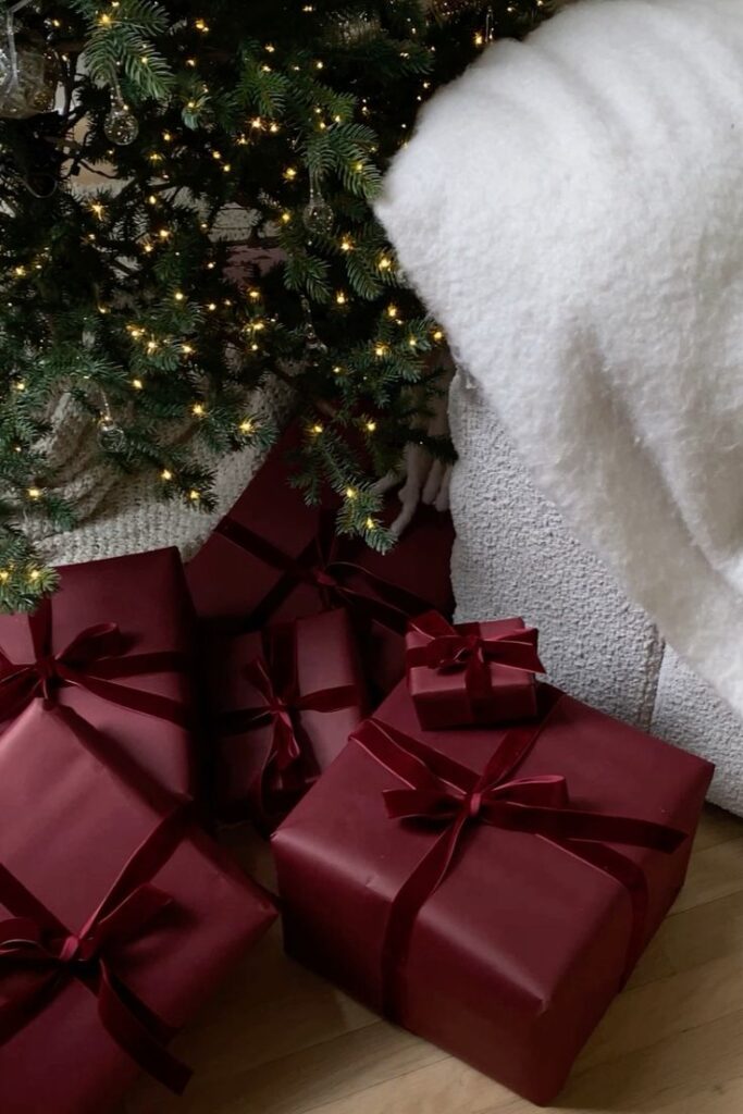 Christmas gifts in red wrapping under decorated tree with glowing lights and cozy white blanket nearby.