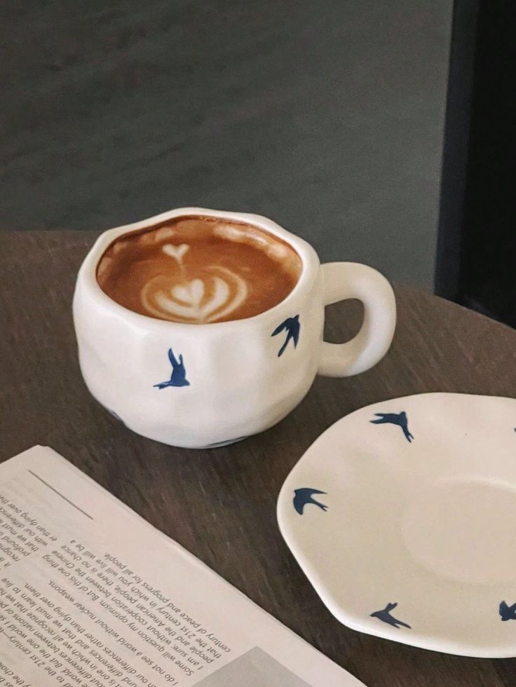 Ceramic cup of latte with bird design on wooden table, next to a matching saucer and open book. Cozy coffee break ambiance.