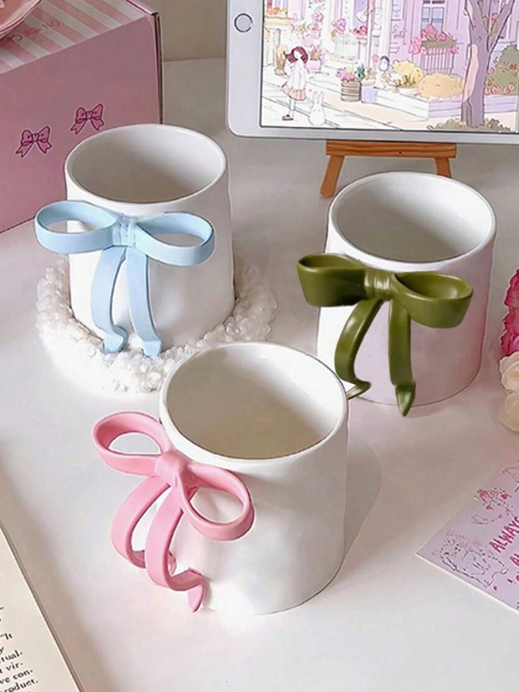 Three elegant mugs with colorful bow handles: pink, blue, and green, on a table near a tablet displaying art.