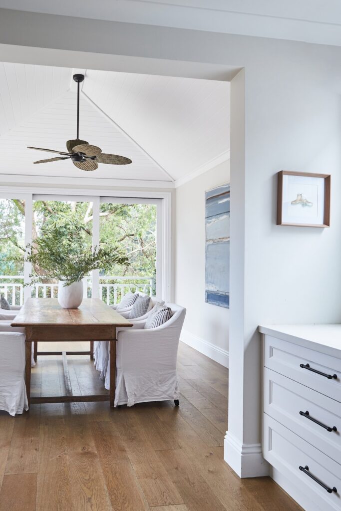 Bright dining area with wooden table, white chairs, large windows, and ceiling fan; contemporary interior design.