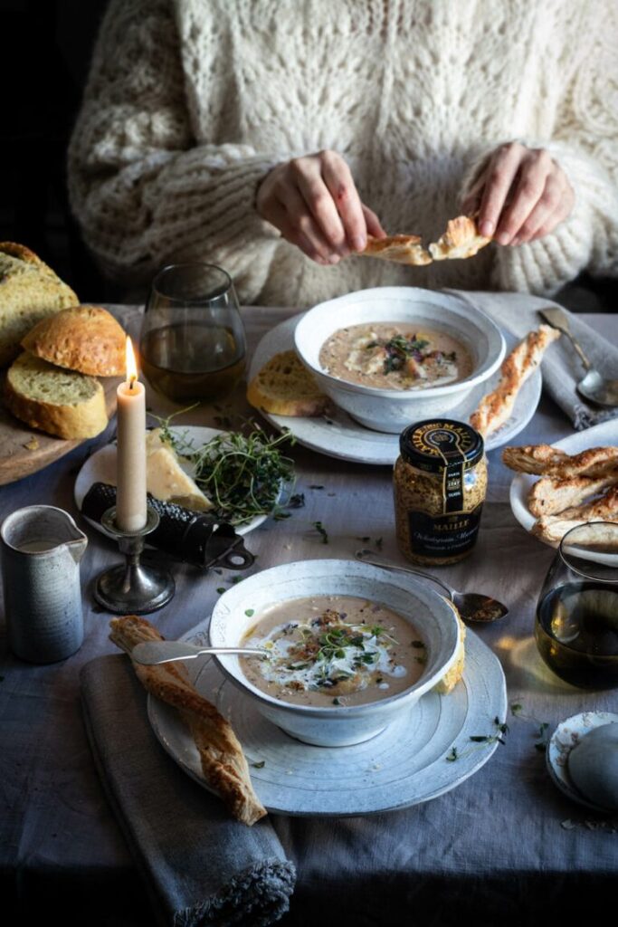 Cozy dinner scene with creamy soup, bread, candlelight, and herbs on a rustic table.