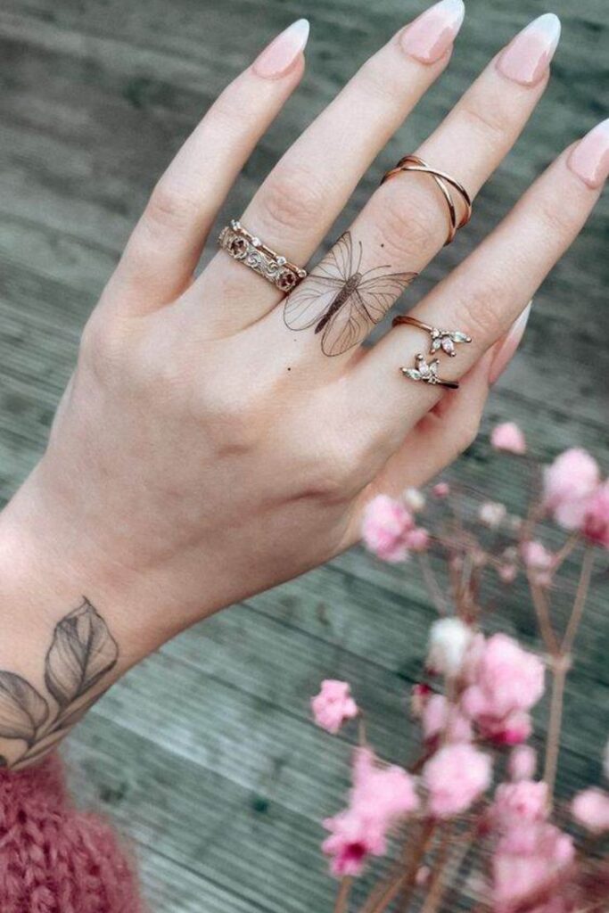 Hand with butterfly tattoo, gold rings, and floral nail art, surrounded by pink flowers against a wooden background.